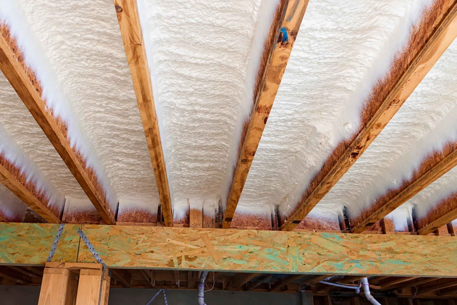 White spray foam fills the spaces between exposed wooden rafters above a plywood beam.