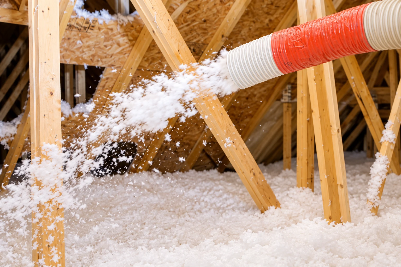 A hose blows loose white insulation across the floor of a timber-framed attic.