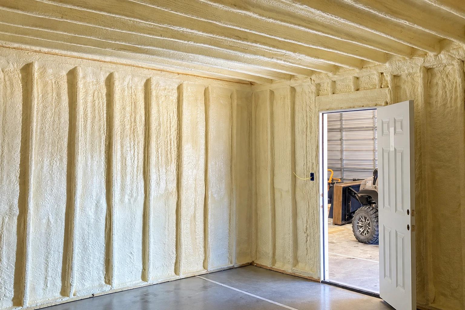 Commercial spray foam insulation coats the walls and ceiling of a garage, beside an open door.