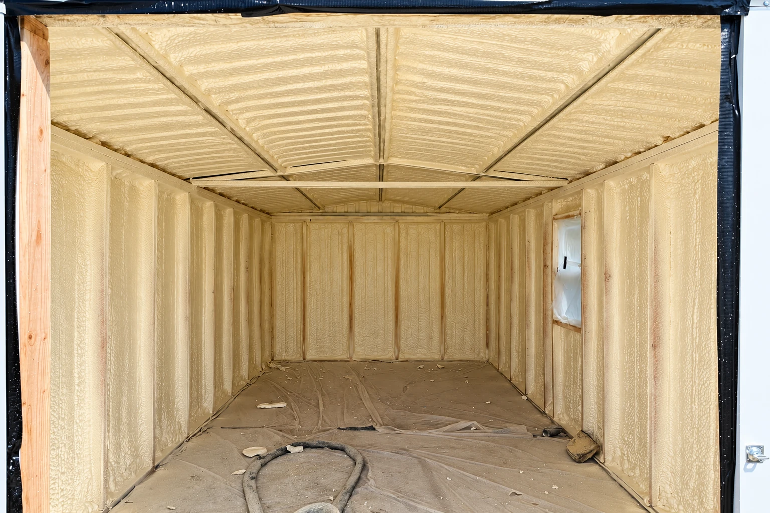 A commercial structure is coated inside with pale yellow spray foam across its walls and ceiling, with a covered floor and hose.