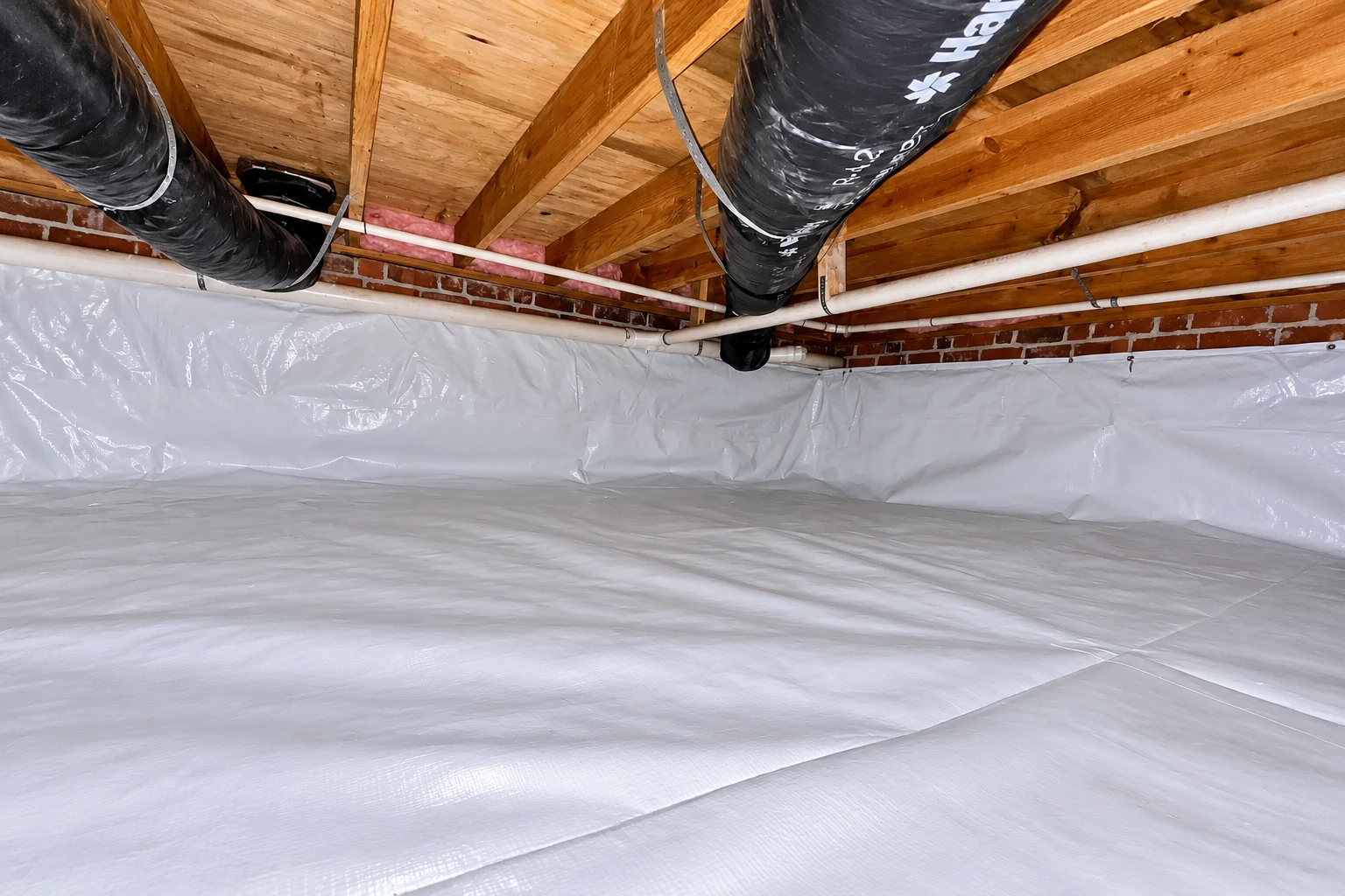 White vapor barrier covers the floor and lower walls of a crawl space beneath exposed joists, ducts, and pipes.
