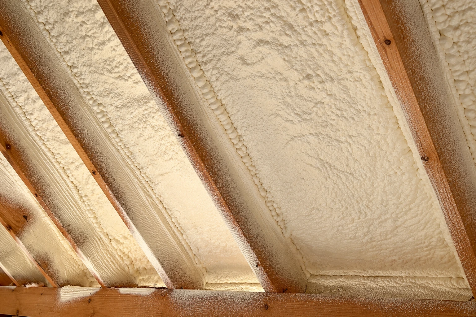 Thick cream-colored spray foam fills the spaces between wooden rafters.