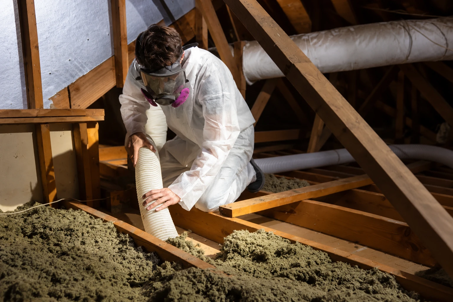 A worker in protective coveralls and a respirator uses a hose to remove loose attic insulation.