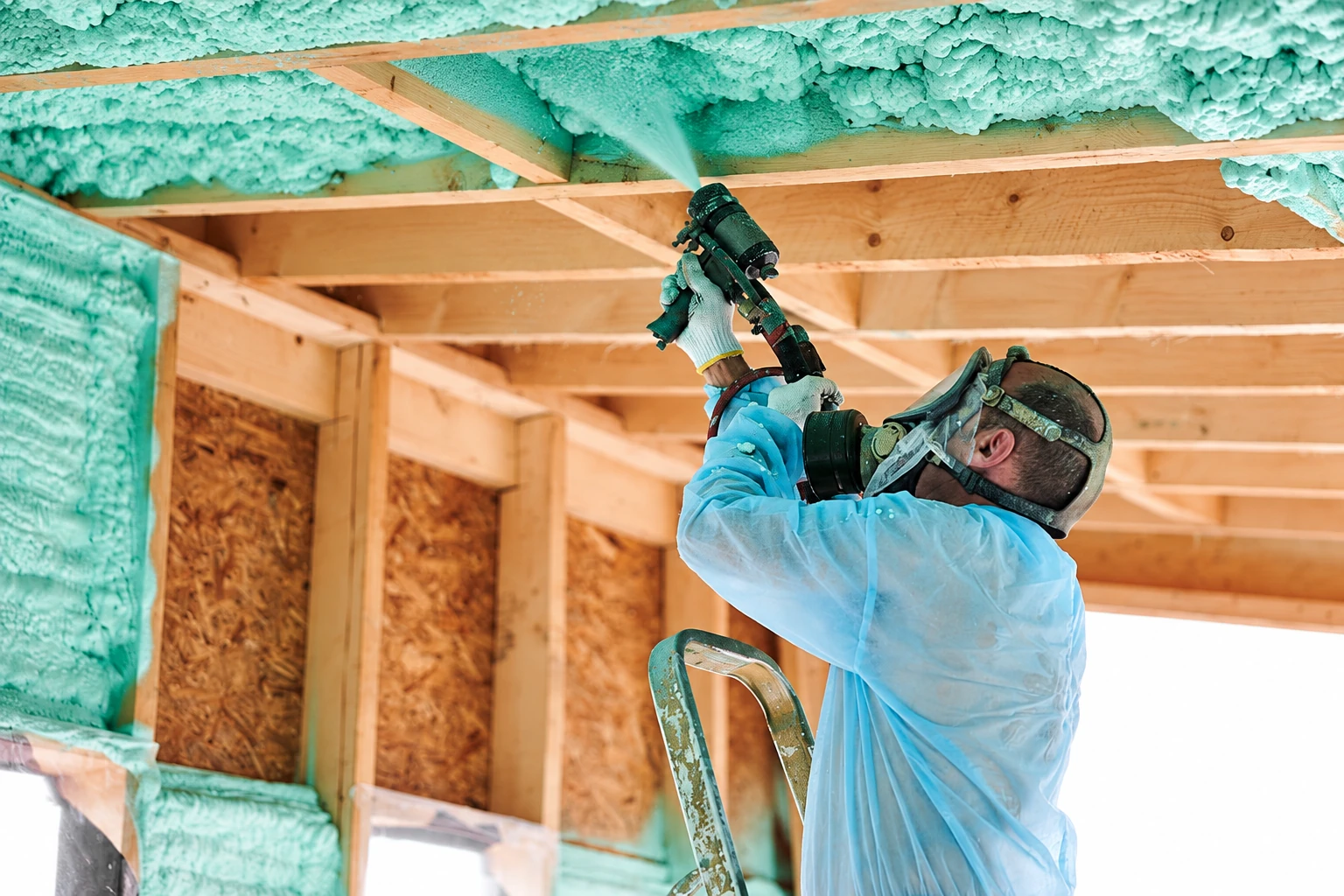 A worker in protective gear sprays mint-green open-cell foam onto the underside of a wooden ceiling.