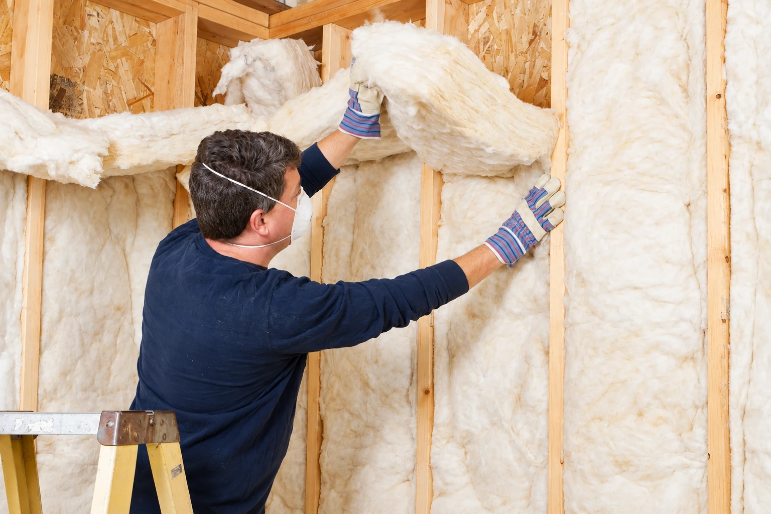 A worker in a mask and gloves fits fluffy white insulation between wooden wall studs.