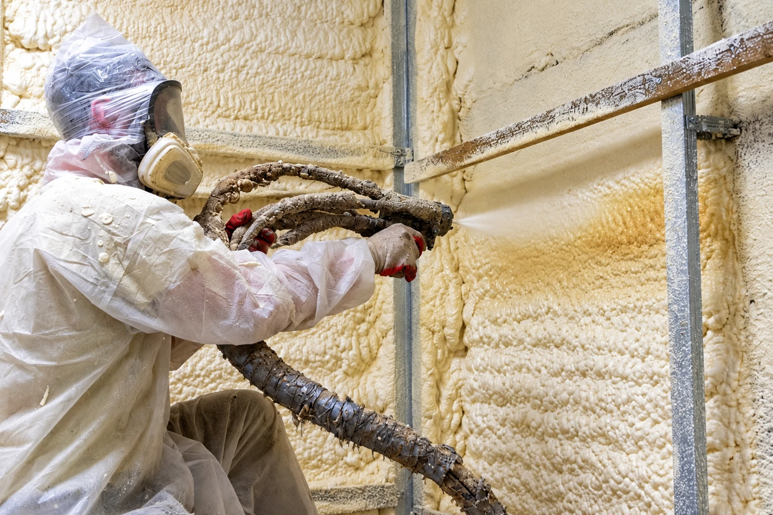 A worker in protective gear sprays pale foam insulation onto a framed wall with a hose.