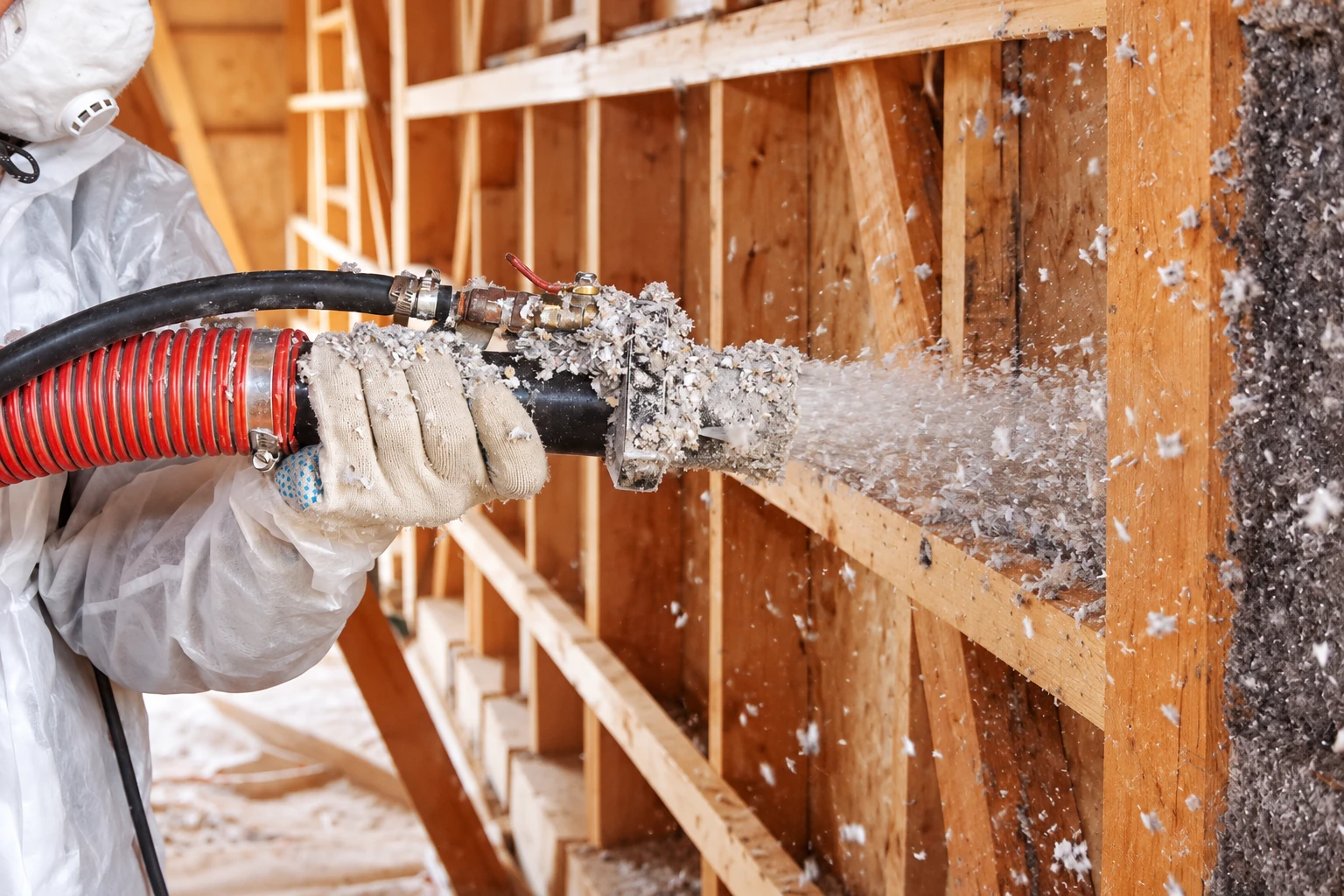 Worker in protective gear sprays loose-fill insulation from a hose into an open wall cavity framed with wood studs.